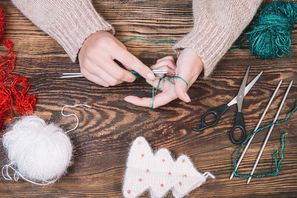 Main de femme qui tricote au crochet des décorations de Noël avec de la laine blanche, de la laine rouge et de la laine verte.
