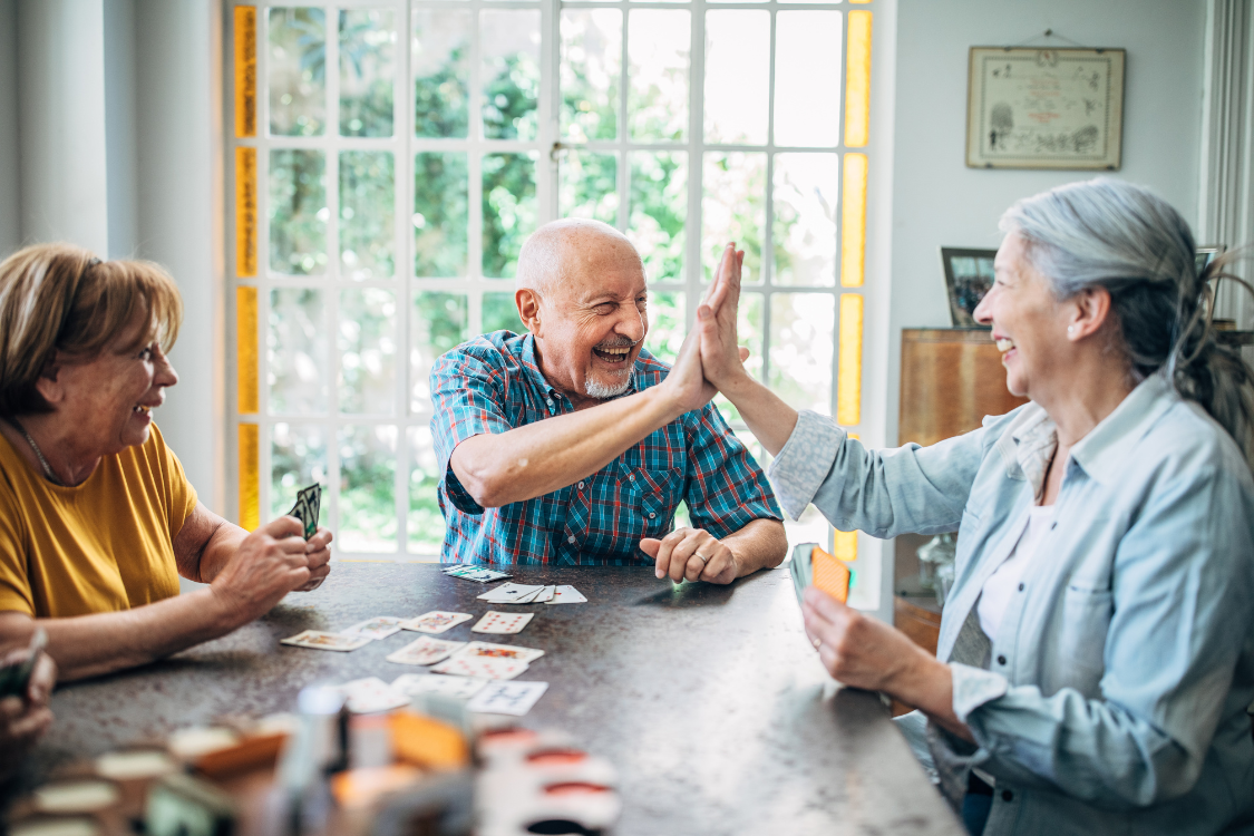 Trois personnes âgées assises autour d'une table en train de jouer aux cartes. Deux d'entre elles échangent un geste de « high five » avec des expressions joyeuses, témoignant d'un moment de convivialité et de plaisir. La pièce est lumineuse grâce à une grande fenêtre en arrière-plan, créant une atmosphère chaleureuse et amicale.