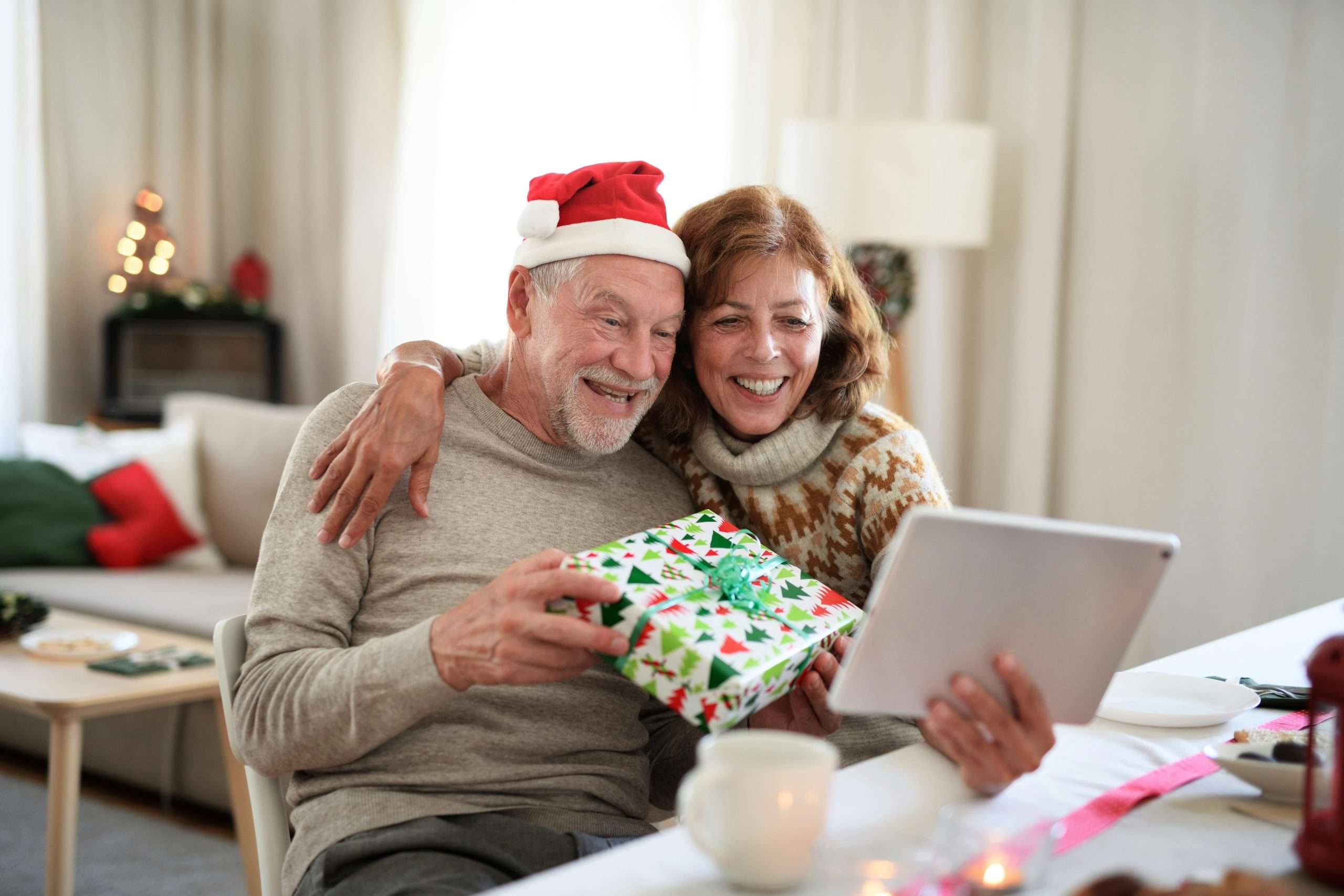 Un couple de personnes âgées, souriants et enlacés, célèbre Noël à distance en faisant un appel vidéo sur une tablette. L'homme porte un chapeau de Noël et tient un cadeau emballé. L'arrière-plan est décoré de façon festive, avec des coussins rouges et verts et un petit sapin illuminé, créant une ambiance chaleureuse et joyeuse.