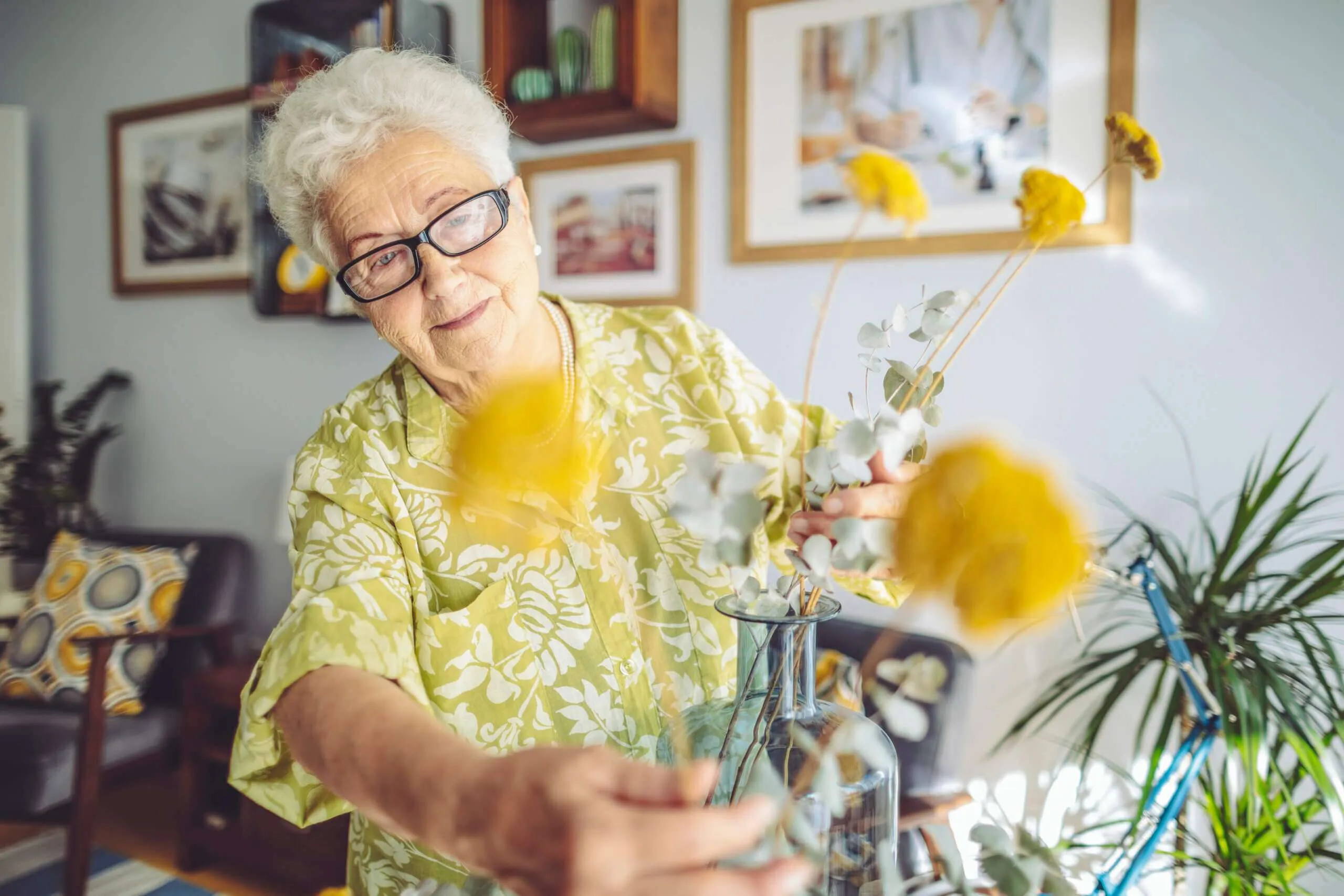Une personne âgée arrange des fleurs jaunes dans un vase en verre bleu, dans un intérieur décoré de plantes et de tableaux.
